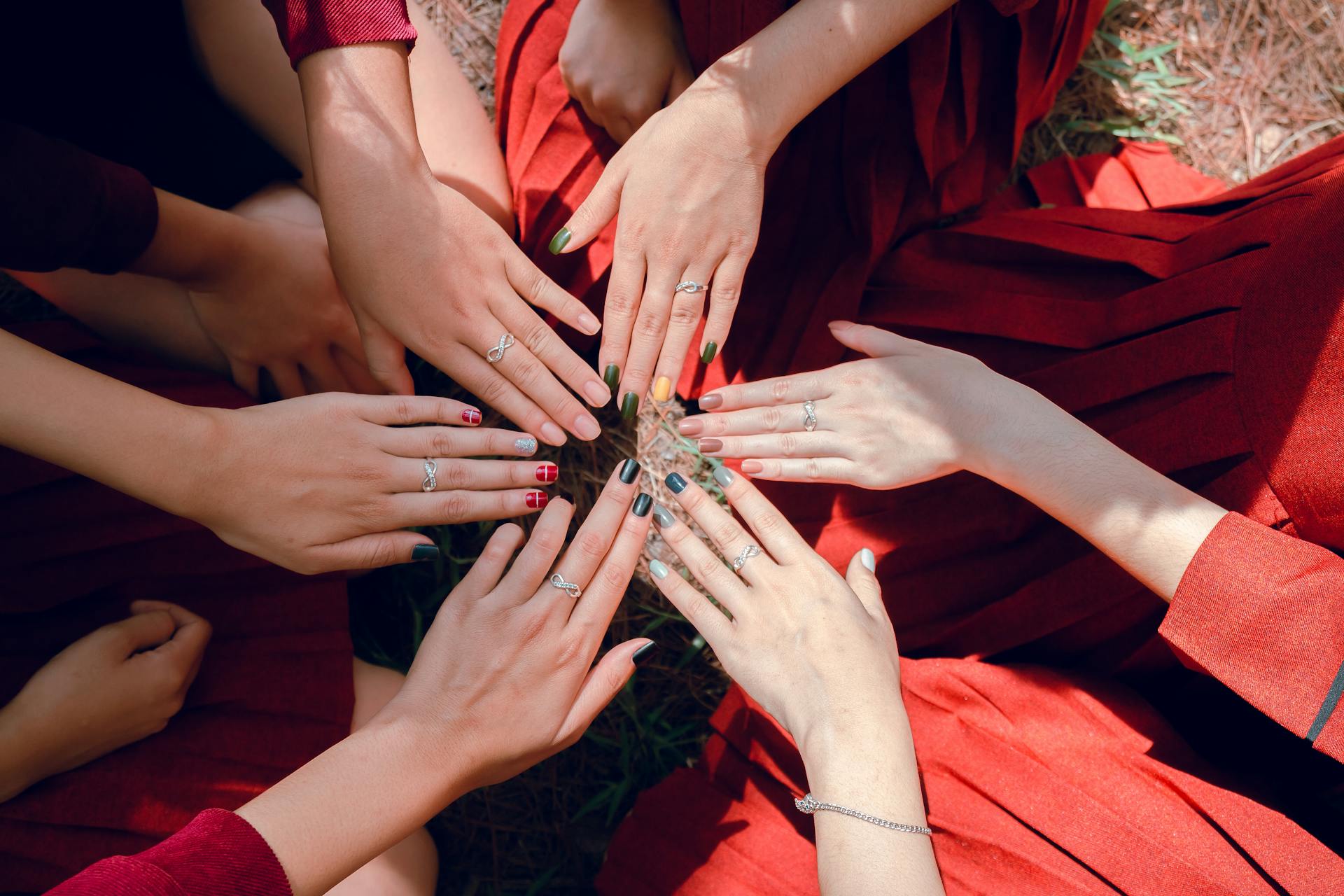 Grupa kobiet siedzących w kręgu, z rękami wyciągniętymi ku środkowi, pokazujących kolorowe paznokcie i pierścionki